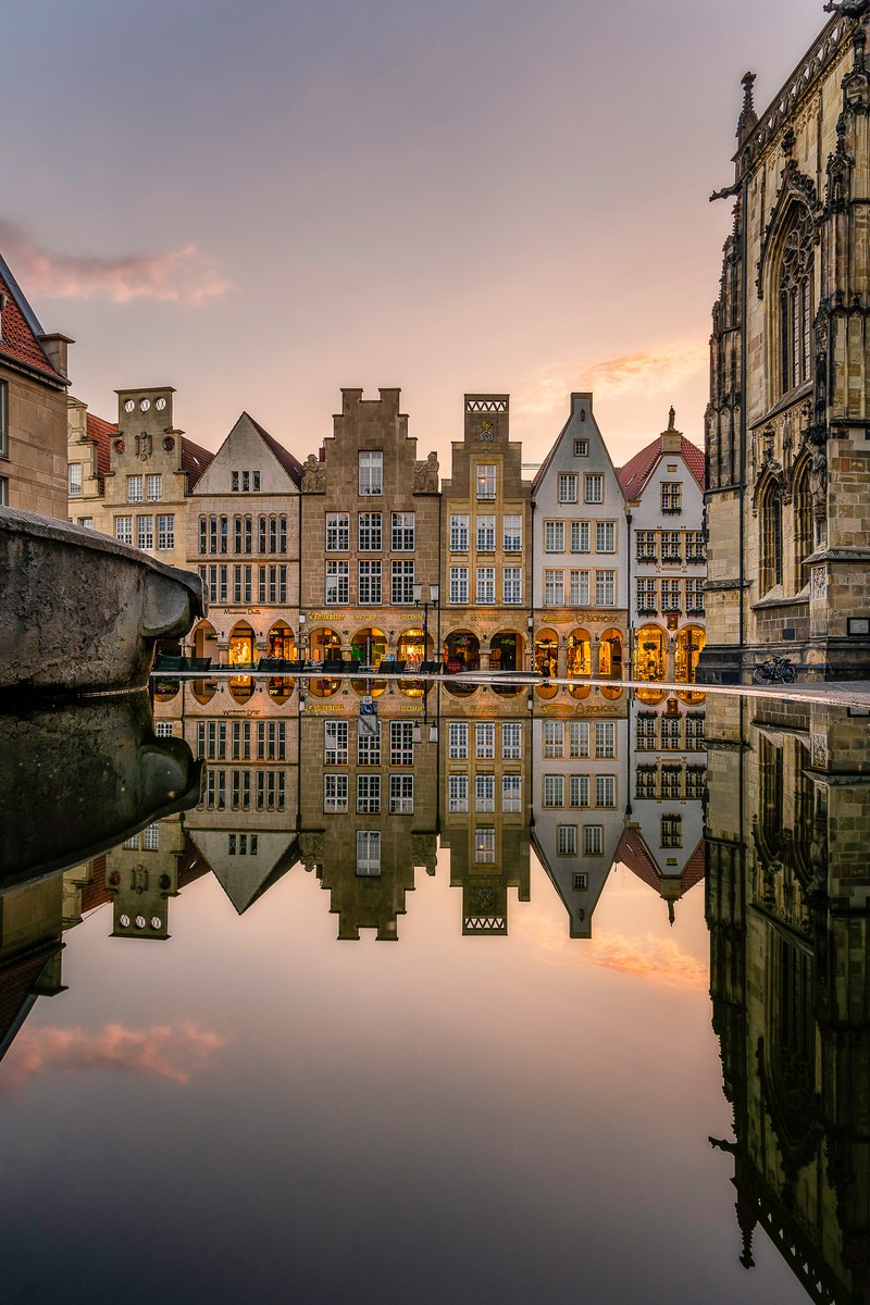 Lambertibrunnen am Prinzipalmarkt in Münster im Abendlicht – die giebelständigen Bürgerhäuser spiegeln sich im Wasser der Brunnenschale, rechts die gotische St.-Lamberti-Kirche. Foto © Sascha Talke.