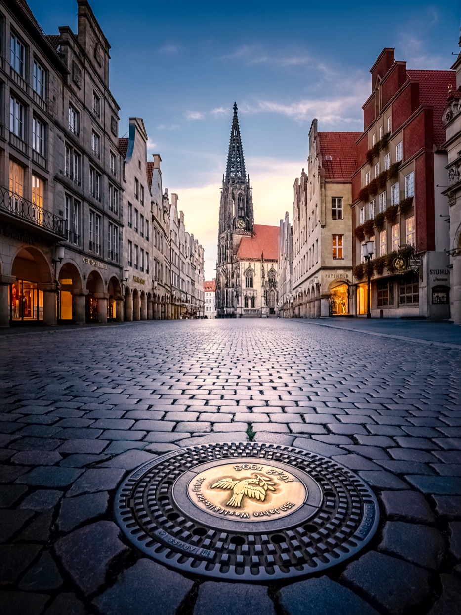 St.-Lamberti-Kirche Münster am frühen Morgen – filigraner Turm über dem leeren Prinzipalmarkt, Kopfsteinpflaster mit Kanaldeckel im Vordergrund. Foto © Thomas Branse.