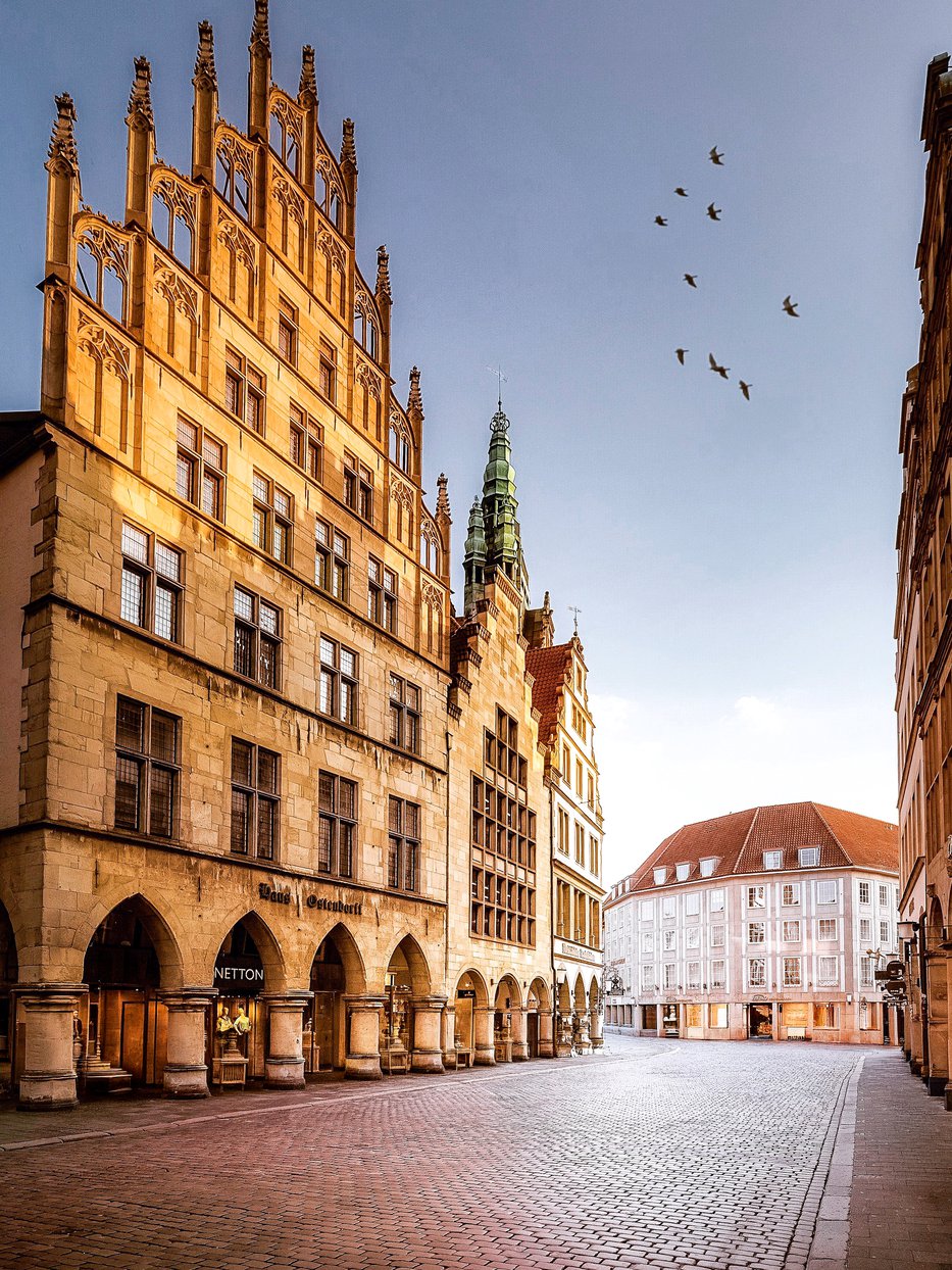 Historisches Rathaus Münster am frühen Morgen – gotische Giebelfassade im Gegenlicht, leerer Prinzipalmarkt, Turm der Lambertikirche im Hintergrund. Foto © Thomas Branse.