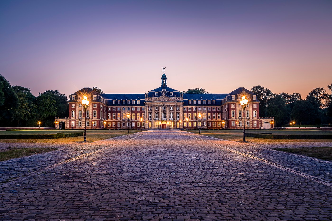 Das Fürstbischöfliche Schloss in Münster frontal zur blauen Stunde – die symmetrische barocke Dreiflügelanlage aus rotem Backstein und hellem Baumberger Sandstein, warm beleuchtete Fenster und Laternen, die Kopfsteinpflaster-Allee führt mittig auf das Schloss zu. Foto © Sascha Talke.